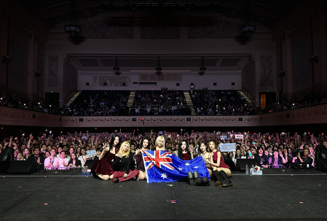 A group of performers sitting on stage, holding an Australian flag, with a large audience cheering in the background.
