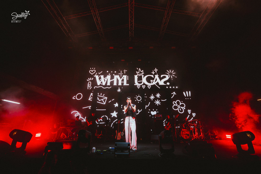 A vocalist performing on stage with a vibrant light show, featuring a large backdrop displaying the text 'WHY LUGA?' amidst colorful graphic doodles and red stage lighting.