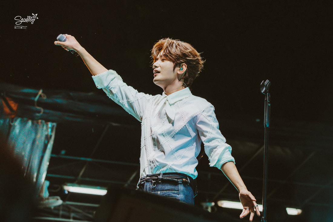 A performer on stage reaches out with a microphone in hand, wearing a white shirt and jeans, with a lively expression and wavy hair.