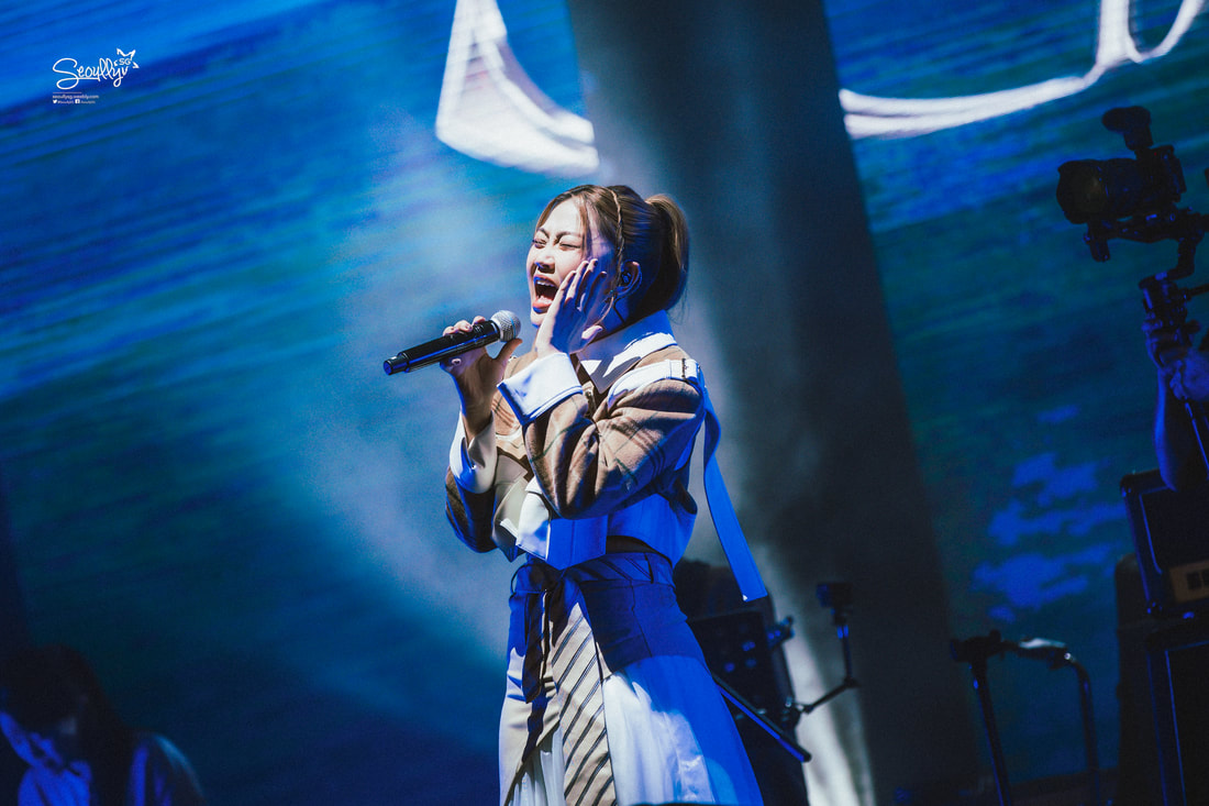 A solo artist passionately singing on stage while holding a microphone, dressed in a stylish outfit, with dramatic lighting and a vibrant background.