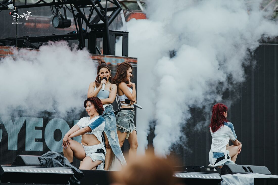 A group of four female dancers performing on stage, surrounded by smoke effects, with one singer holding a microphone and the others dancing energetically.