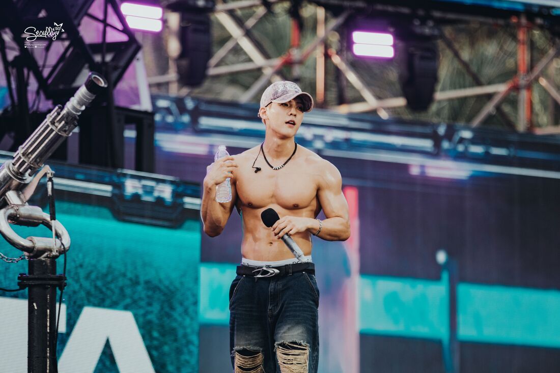 A shirtless male performer on stage, holding a microphone and a water bottle, with a rugged denim look, against a colorful backdrop of lights and stage equipment.