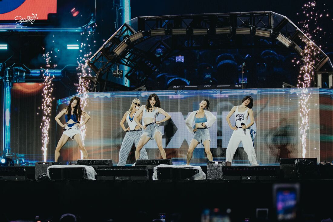 A group of five female performers dancing on stage during a live concert, with colorful stage lights and fireworks in the background.