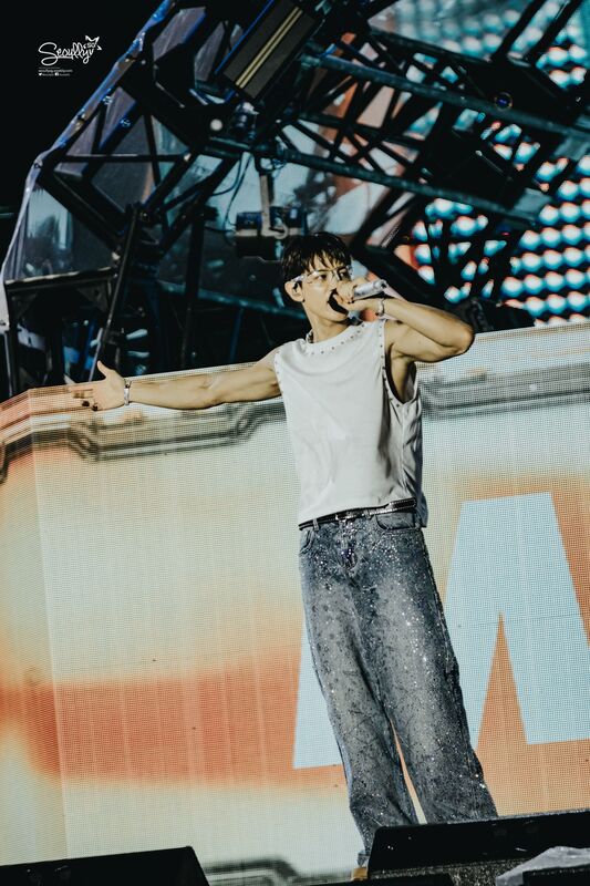 A performer on stage with short hair, wearing a sleeveless white top and sparkly gray pants, holding a microphone and gesturing with one arm while singing.