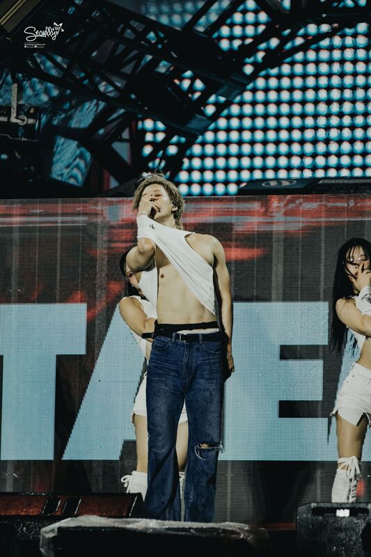 A male performer wearing a sleeveless shirt and ripped jeans passionately singing on stage, with dancers behind him and a bright LED screen in the background.