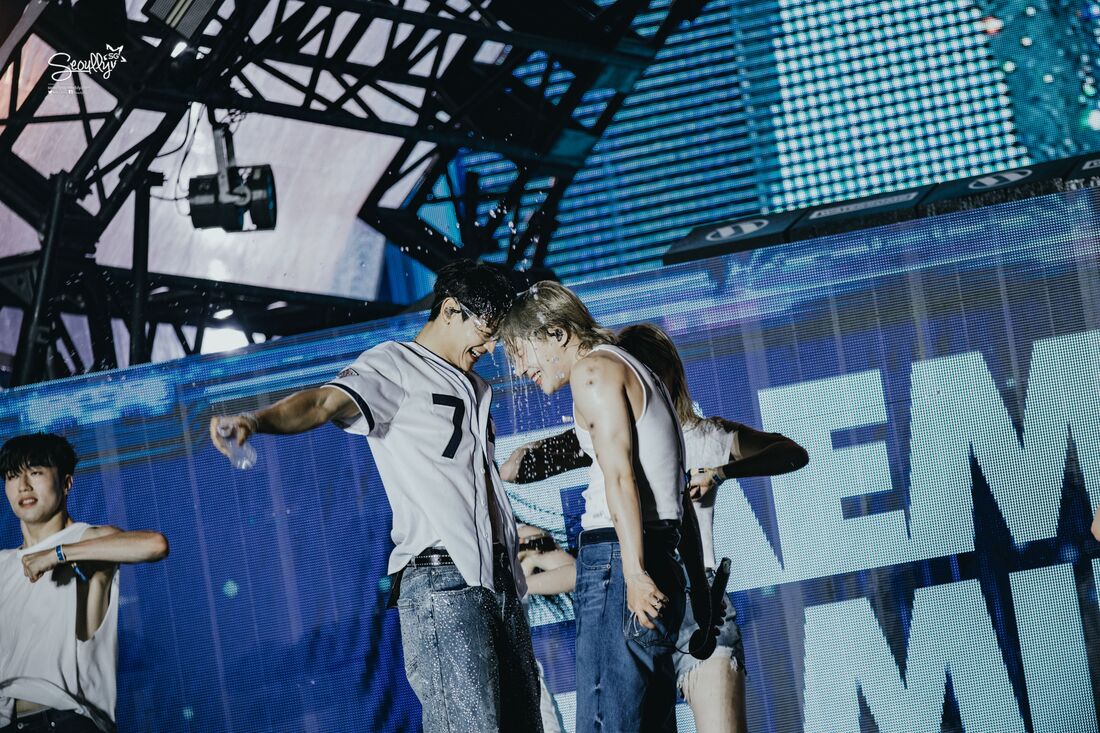 Two male performers on stage with water splashing around them, both wearing casual clothing. One is holding a water bottle while smiling at the other, who is wearing a white tank top. A female performer is partially visible behind them.