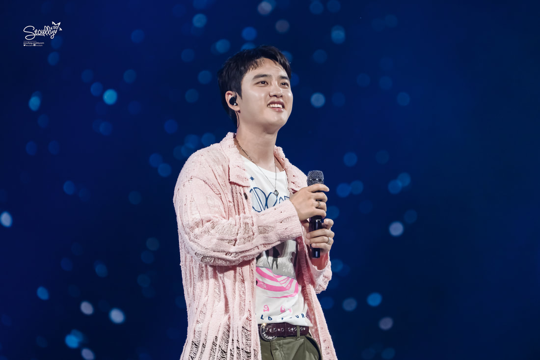 A young man smiling and singing on stage, wearing a pink sweater and a graphic t-shirt, holding a microphone, with a blurred blue background.