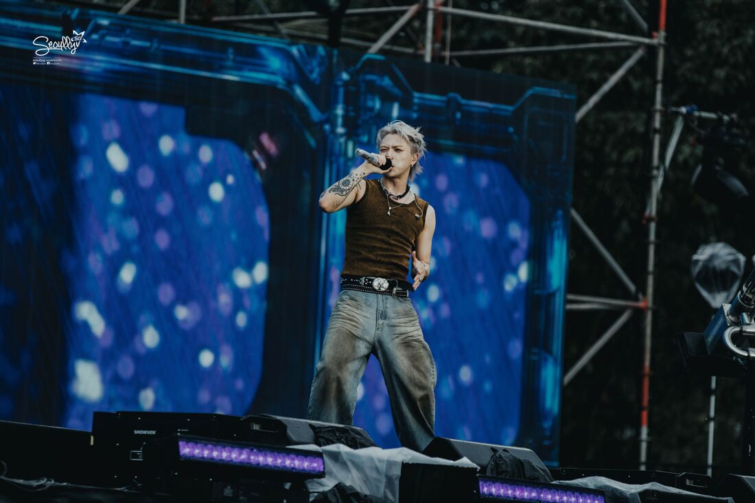 A performer singing on stage with a microphone, wearing a brown sleeveless shirt and loose gray pants, with a vibrant backdrop of blue and purple lights.