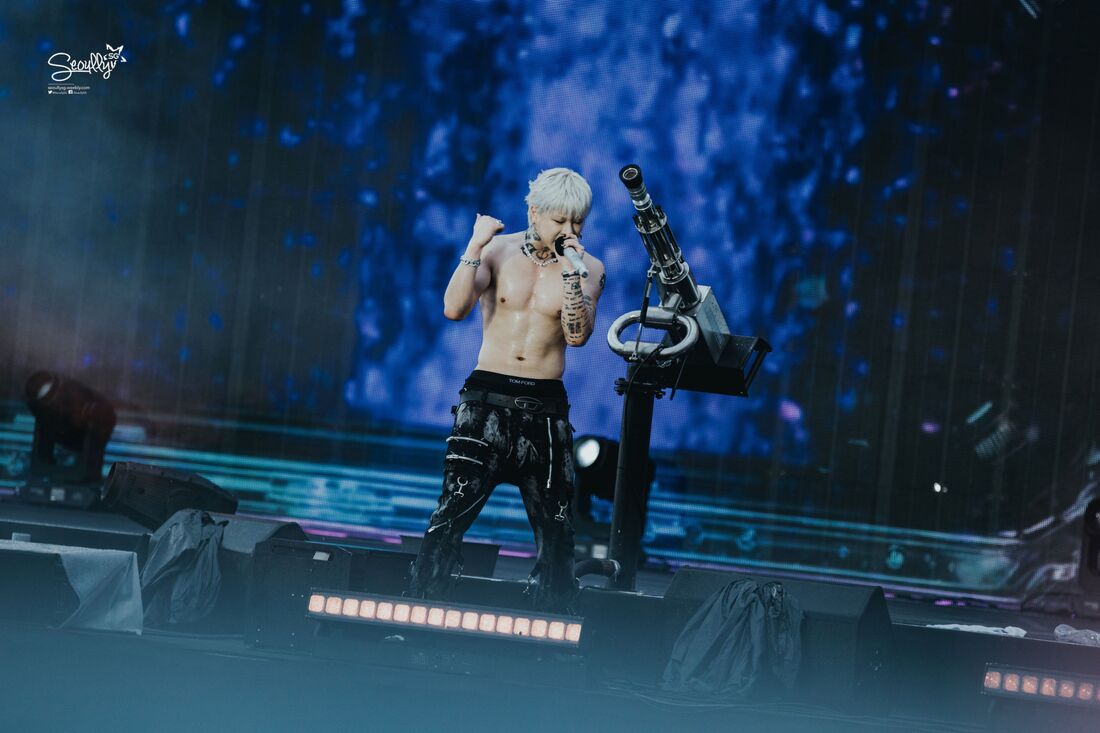 A shirtless performer with platinum blonde hair sings on stage, surrounded by a colorful light display. He is wearing black pants with various designs and appears energetic.