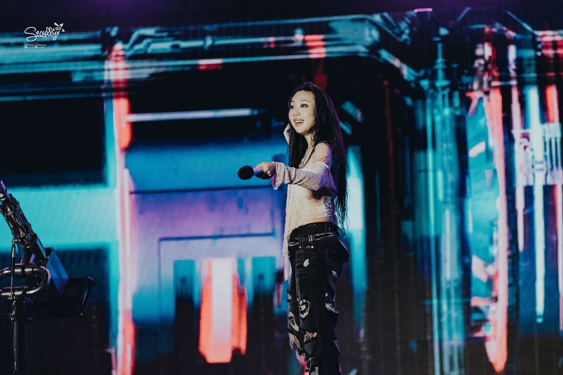 A performer on stage holds a microphone, smiling and engaging with the audience. She is wearing a light, stylish top and distressed black jeans, with long hair cascading down her shoulders. The background features vibrant, colorful stage lights and graphics.