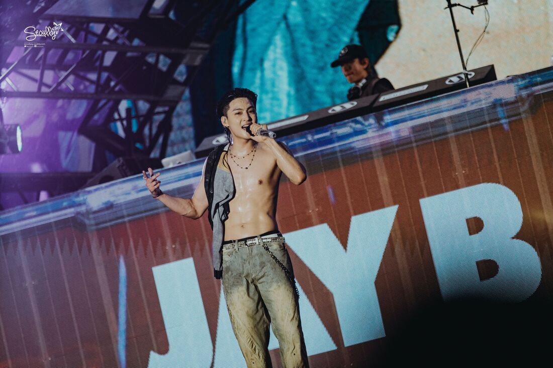 A male performer singing on stage, wearing a tank top and showing his toned torso, with a DJ in the background. The backdrop features colorful lights and a large screen displaying the name 'JAY B'.