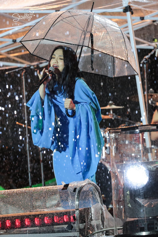A singer performing on stage under a transparent umbrella while it is raining, wearing a blue outfit.