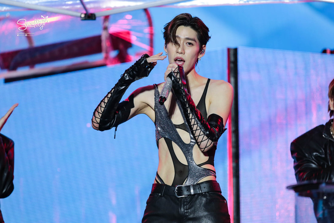 A male performer wearing a stylish, revealing outfit with lace-up details, singing on stage with dramatic lighting in the background.