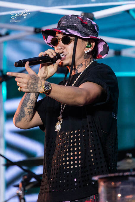 A male performer wearing sunglasses and a patterned bucket hat, passionately singing into a microphone on stage, with tattoos visible on his arms.