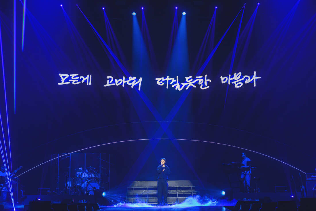 A singer performing on stage with blue lighting and laser beams, while a large text is displayed in the background.