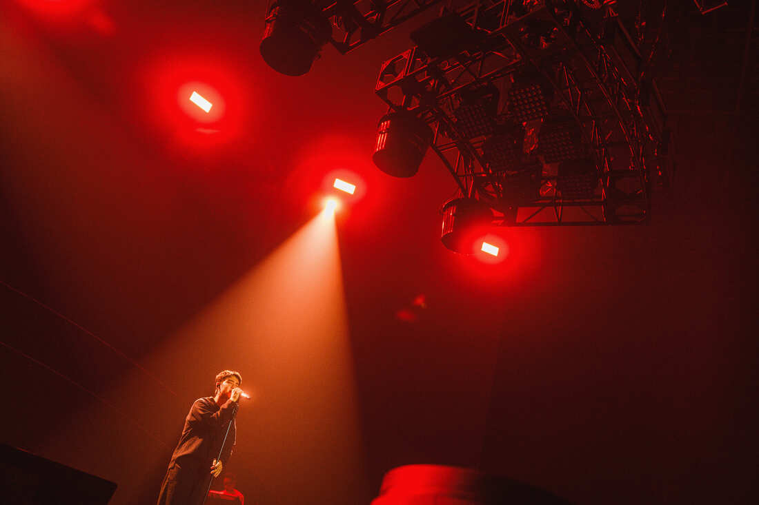 A performer on stage singing under red stage lights, with dramatic lighting from above.