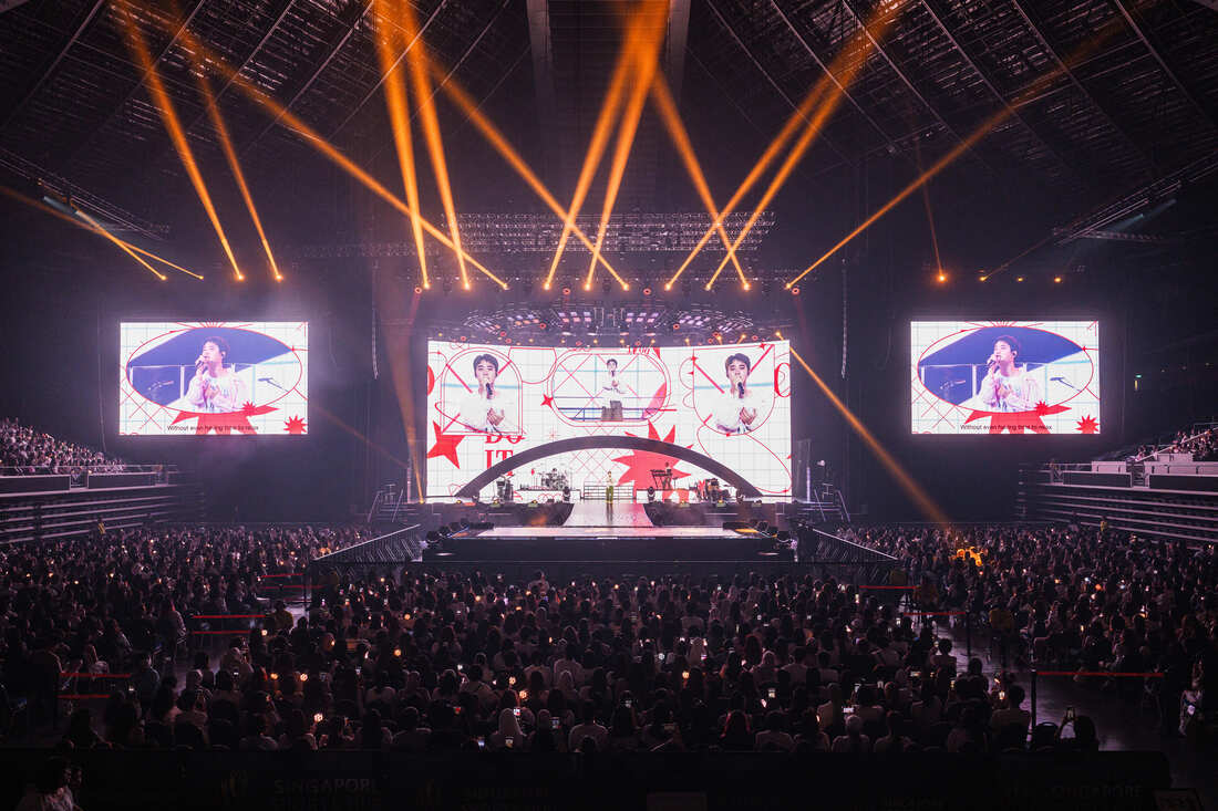 A large crowd enjoys a live music performance on stage, featuring bright stage lights and multiple video screens displaying a performer, in a concert setting.