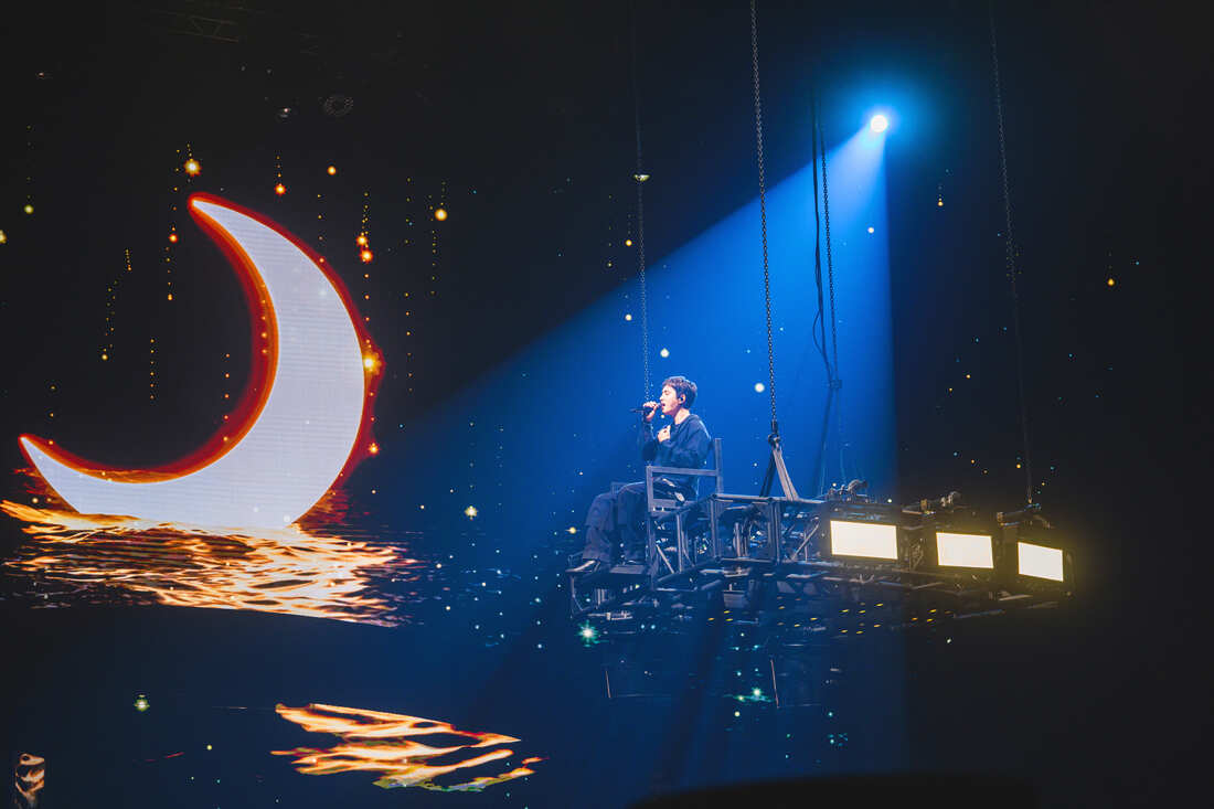 A singer performing on a suspended platform with a crescent moon projection and a starry background.