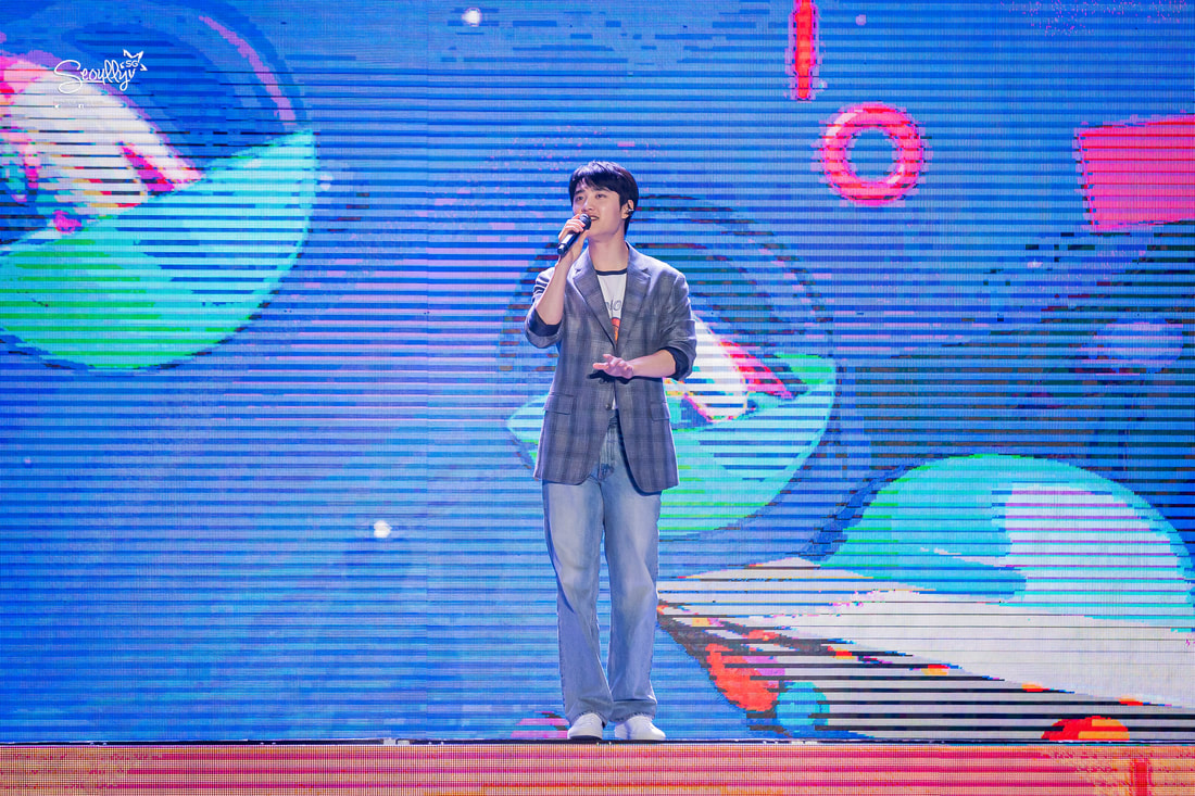 A performer singing on stage, wearing a plaid blazer and casual jeans, with colorful background visuals featuring abstract shapes.