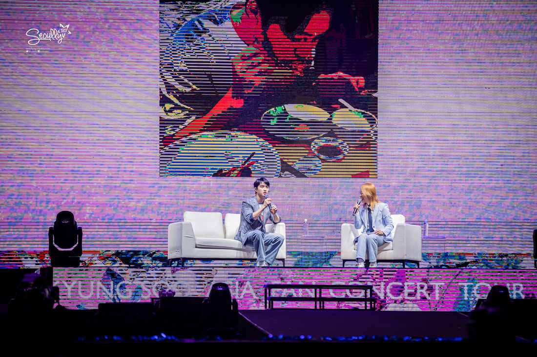 A live event featuring two seated speakers on stage, discussing in front of a large screen displaying a colorful image of a man eating at a table.