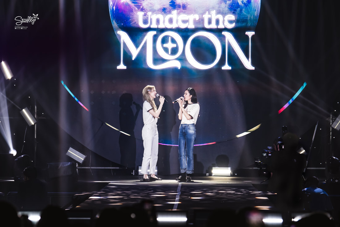 Two female singers performing on stage under a large screen that displays the text 'Under the Moon.' They are holding microphones and engaging with each other while surrounded by stage lights.