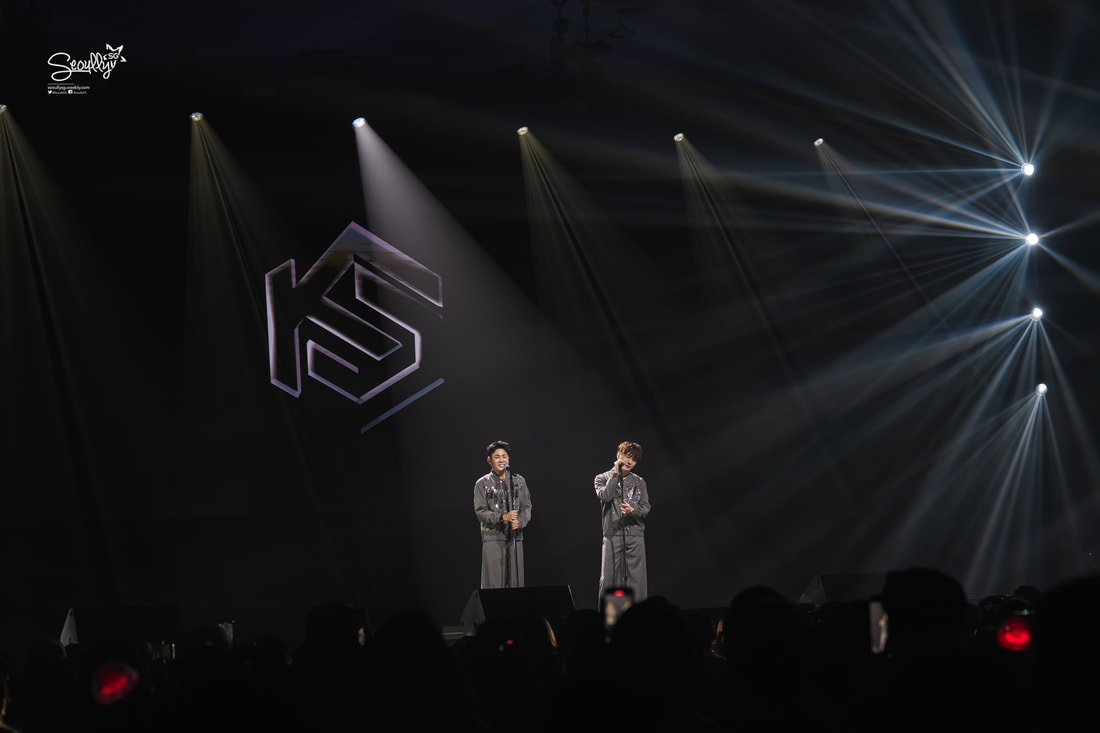 Two performers on stage singing under dramatic lighting at a concert, with a large logo in the background.
