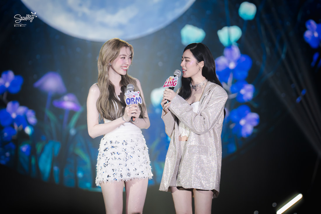 Two women stand on stage with microphones, smiling and engaging with each other. They are surrounded by a colorful backdrop featuring flowers and a moon.
