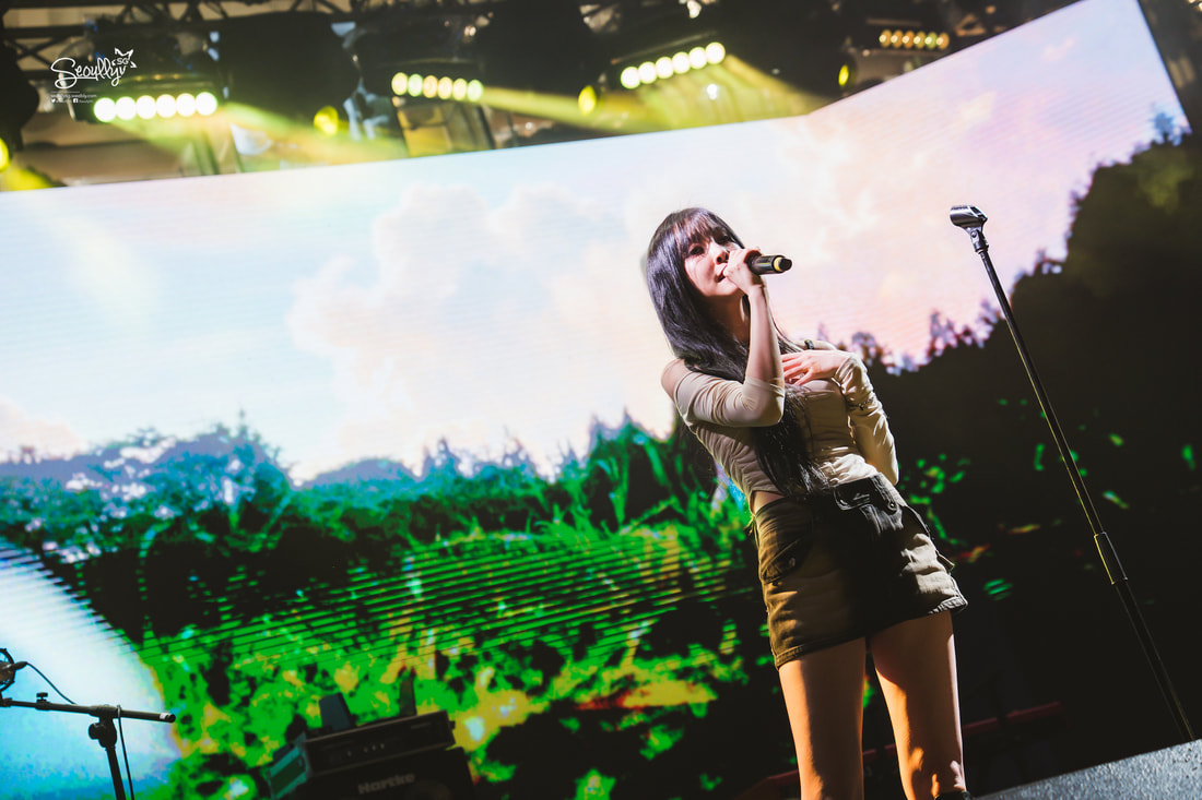 A singer with long black hair performing on stage, holding a microphone. The background features a vibrant display with a nature scene and colorful lights.