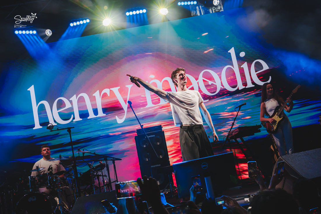 A live performance featuring a male singer on stage, pointing to the audience, with a vibrant backdrop displaying the name 'henry moodie.' In the foreground, a drummer is visible, while a female guitarist performs beside him.