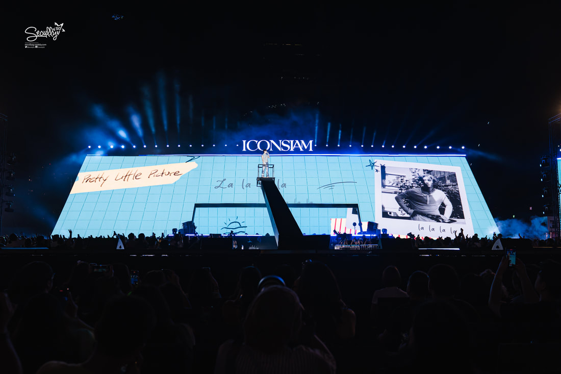 A large stage featuring a vibrant display with the text 'Pretty Little Future' and 'La la la la' in a colorful design, along with a photo of a person. Bright lights and a cheering audience are visible in the foreground at an event at ICONSIAM.