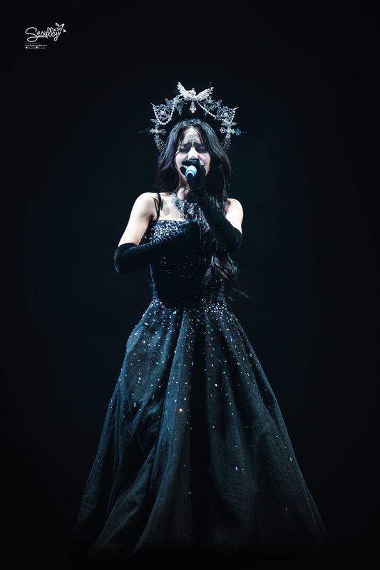 A female singer performs on stage wearing a black gown adorned with sequins, long gloves, and a crown, while holding a microphone against a dark background.
