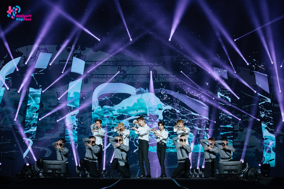 A group of performers dancing on stage under vibrant lights during a live concert at Hallyu Pop Fest, with a digital screen in the background displaying colorful visual effects.