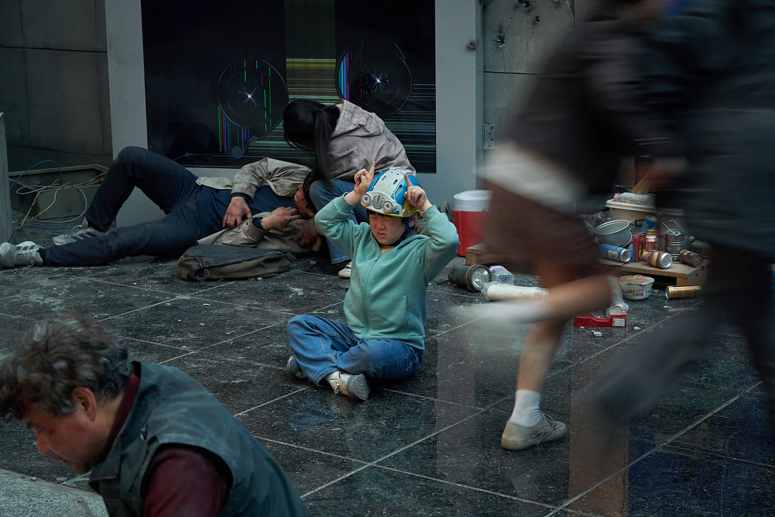 A young child wearing a helmet sits on the ground, appearing distressed, while people move quickly in the background, some lying on the ground amidst scattered trash and debris.
