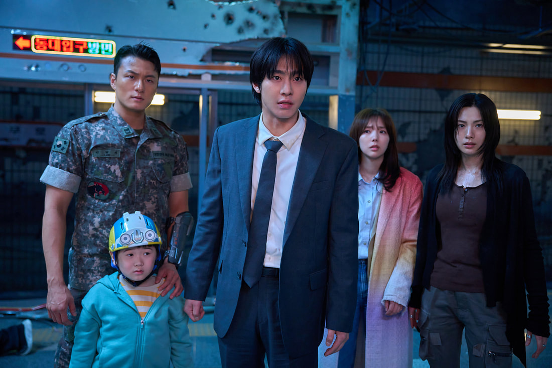 A group of five individuals stands together in a dimly lit urban environment. They display a range of expressions, suggesting tension or concern. The group includes a soldier in military attire, a man in a suit, a child wearing a helmet and jacket, and two women, one in a long colorful coat and the other in casual wear.