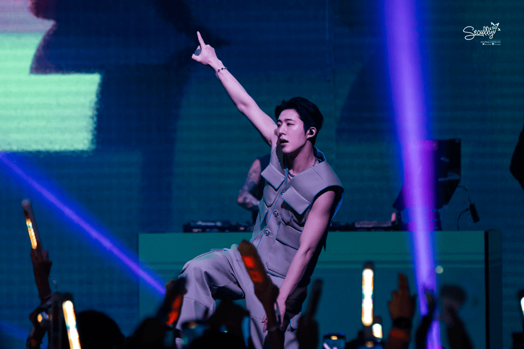A male performer on stage wearing a sleeveless outfit, striking a dynamic pose with one arm raised, while fans in the foreground wave light sticks and cheer.