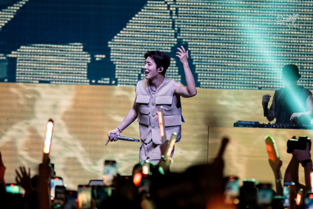 A performer on stage wearing a light-colored outfit, smiling and engaging with the audience. The background features bright lights and screens, while fans hold up their phones to capture the moment.