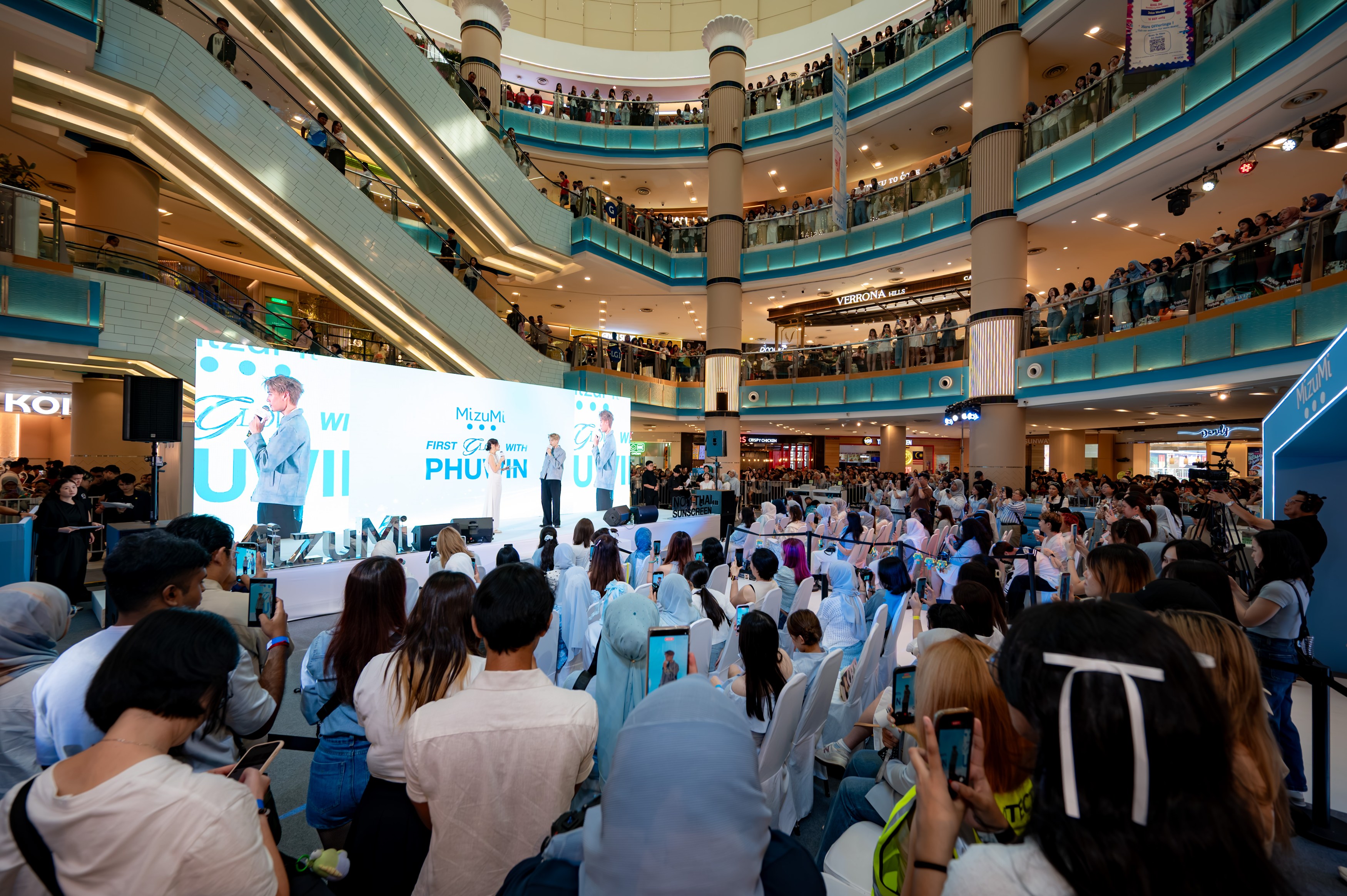 A crowded shopping mall event with a stage featuring a speaker, surrounded by an audience taking pictures with their phones.
