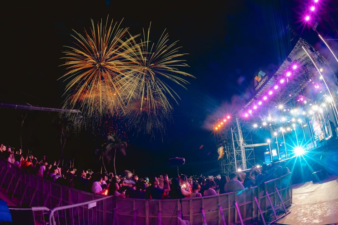 Fireworks display over a concert stage during a nighttime event, with a crowd in the foreground enjoying the show.