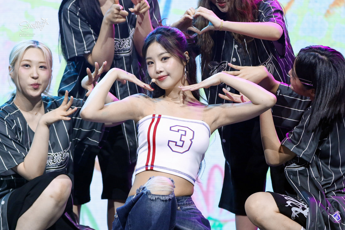 A young female performer striking a pose with her arms raised and head tilted slightly, surrounded by other performers making heart shapes with their hands, all set against a colorful backdrop.