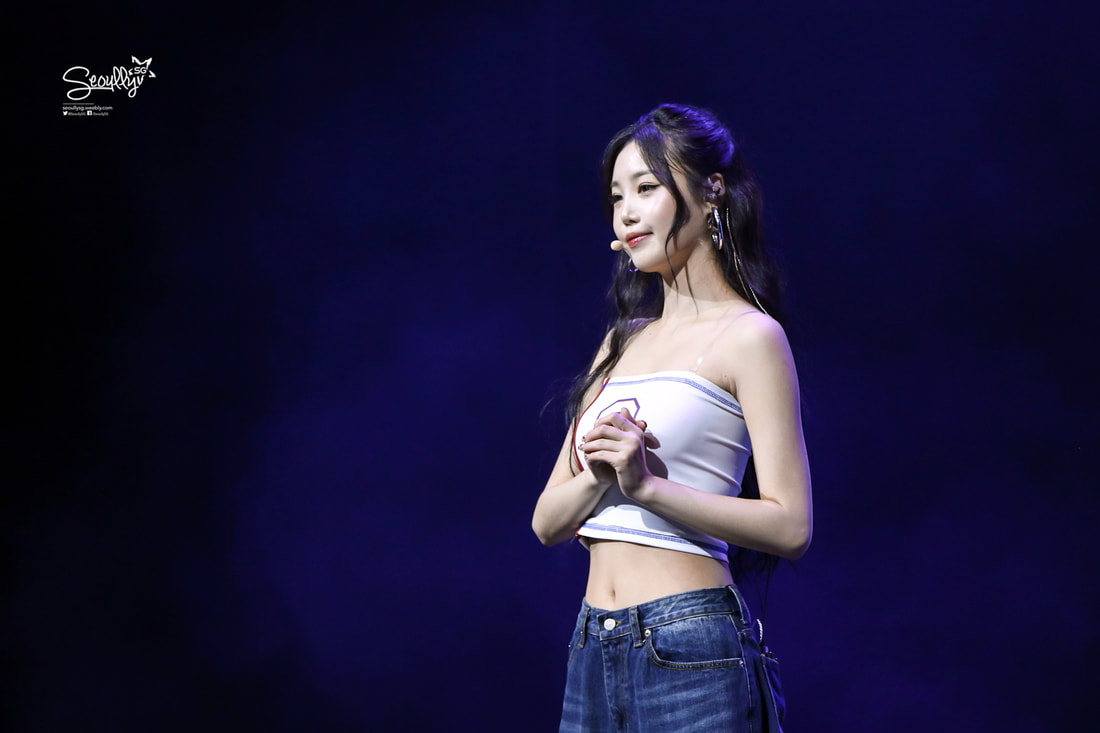 A performer standing on stage with a purple background, wearing a white crop top and blue jeans, with long dark hair and a microphone.