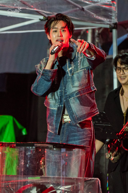 A male performer in a denim jacket and jeans is singing into a microphone on stage, with a cheerful expression. A band member is visible in the background, playing an instrument.