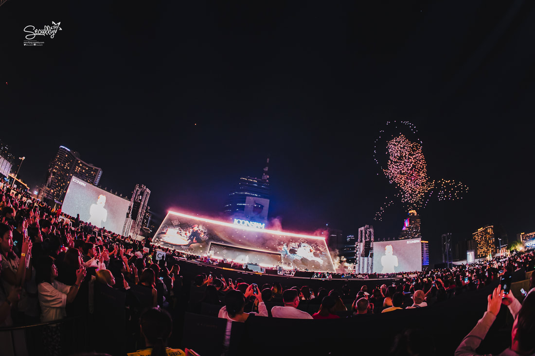 A large crowd is gathered in an outdoor venue at night, watching a performance on big screens. Colorful fireworks and light displays are visible in the sky, creating a festive atmosphere.