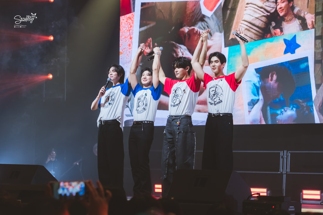 Four performers on stage during a concert, wearing matching graphic t-shirts, posing with arms raised in celebration. A colorful backdrop displays images related to their performance.