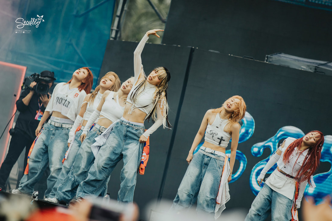 A group of six performers on stage dancing in coordinated outfits, featuring crop tops and loose jeans, during a live performance. A camera crew is visible in the background.