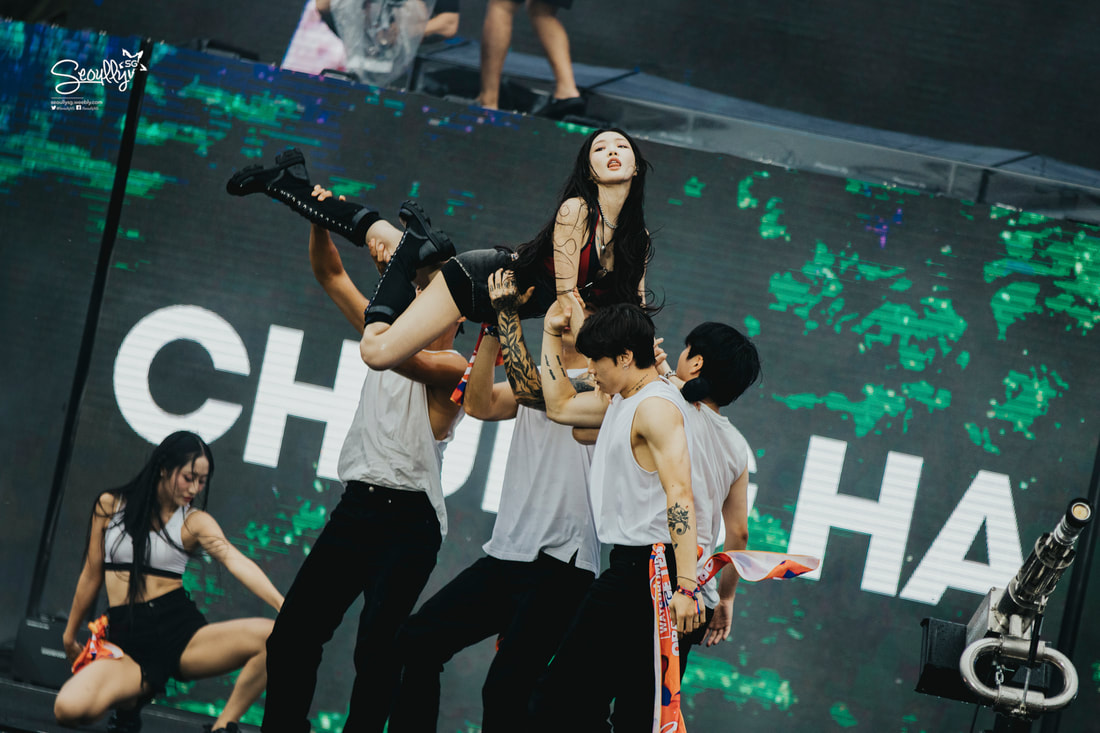 A performer is being lifted by several dancers during a stage performance, with a vibrant backdrop displaying text. The performer wears edgy clothing and has long hair, while the dancers are dressed in casual concert attire.