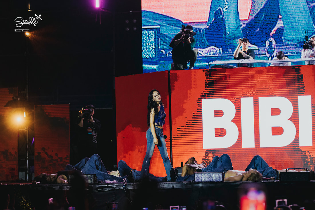 A female performer singing on stage in front of a large screen displaying the name 'BIBI', with several dancers lying on the stage.