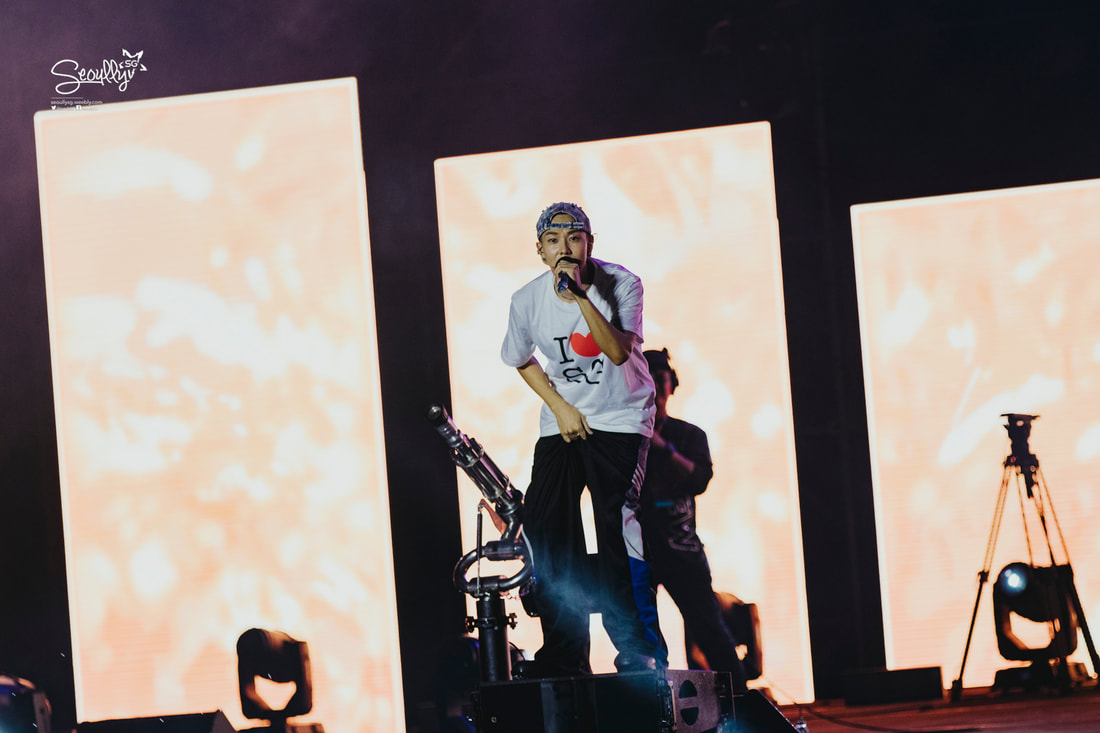 A performer on stage wearing a white t-shirt with a red heart design, holding a microphone and singing, with bright illuminated screens in the background.