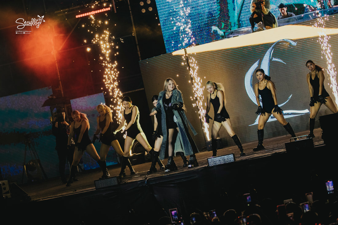 A live performance featuring a female artist in a stylish coat, surrounded by backup dancers in black outfits, with sparklers lighting up the stage and a vibrant lighting display in the background.