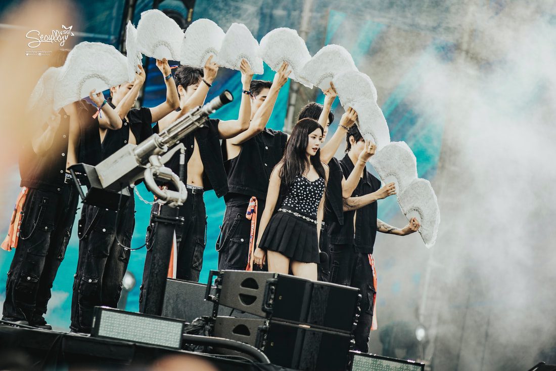 A performer in a black outfit stands in front of background dancers holding large white props during a live performance, with a smoky atmosphere and colorful lighting.