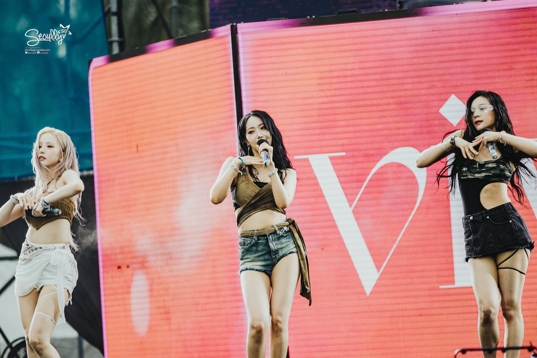 Three female performers on stage dancing and singing, dressed in stylish outfits with a large colorful screen in the background displaying a graphic.
